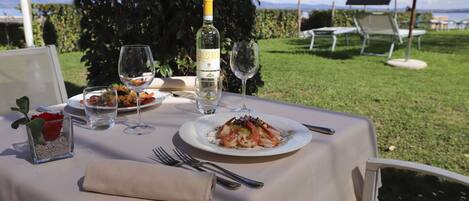 Une table à manger installée à l’extérieur, avec une assiette de nourriture, des verres à vin et une bouteille de vin.