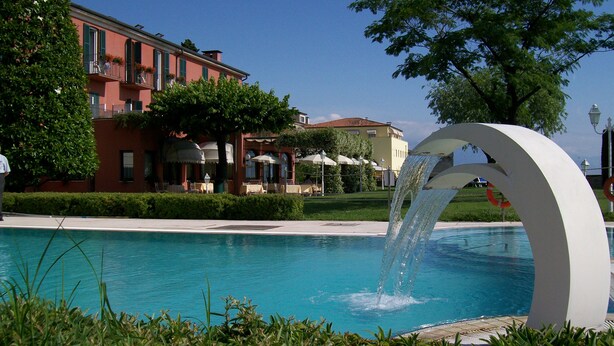 A swimming pool with a water feature, surrounded by buildings and greenery. - Hotel Fonte Boiola (Sirmione)