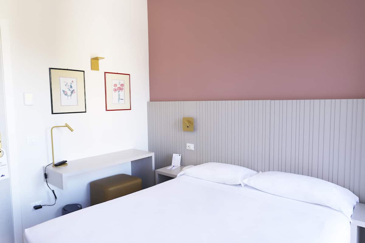 A hotel room with a bed, a desk, a chair, a nightstand, and two framed pictures on the wall.
