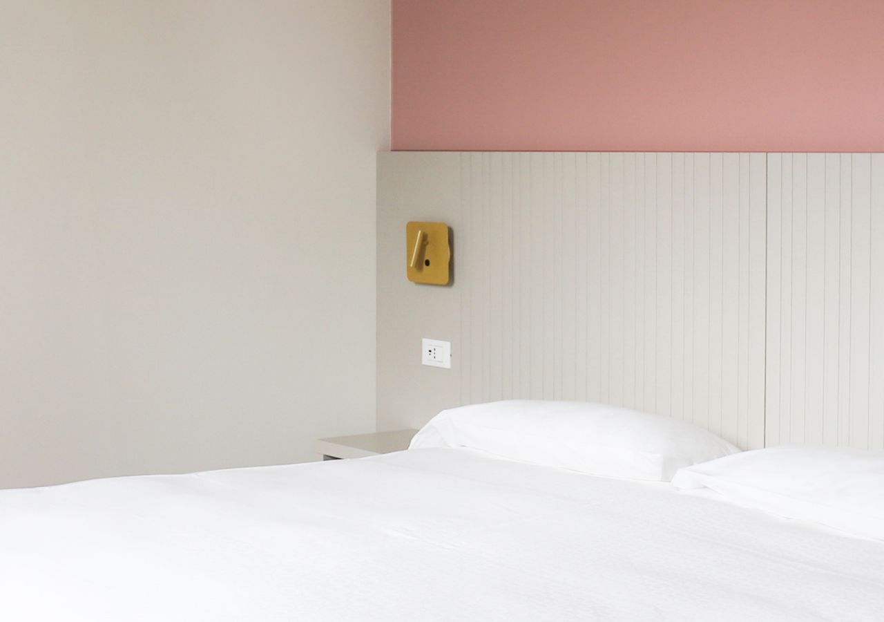 A hotel room with a bed, a nightstand, a wall socket, and a wall-mounted light switch.