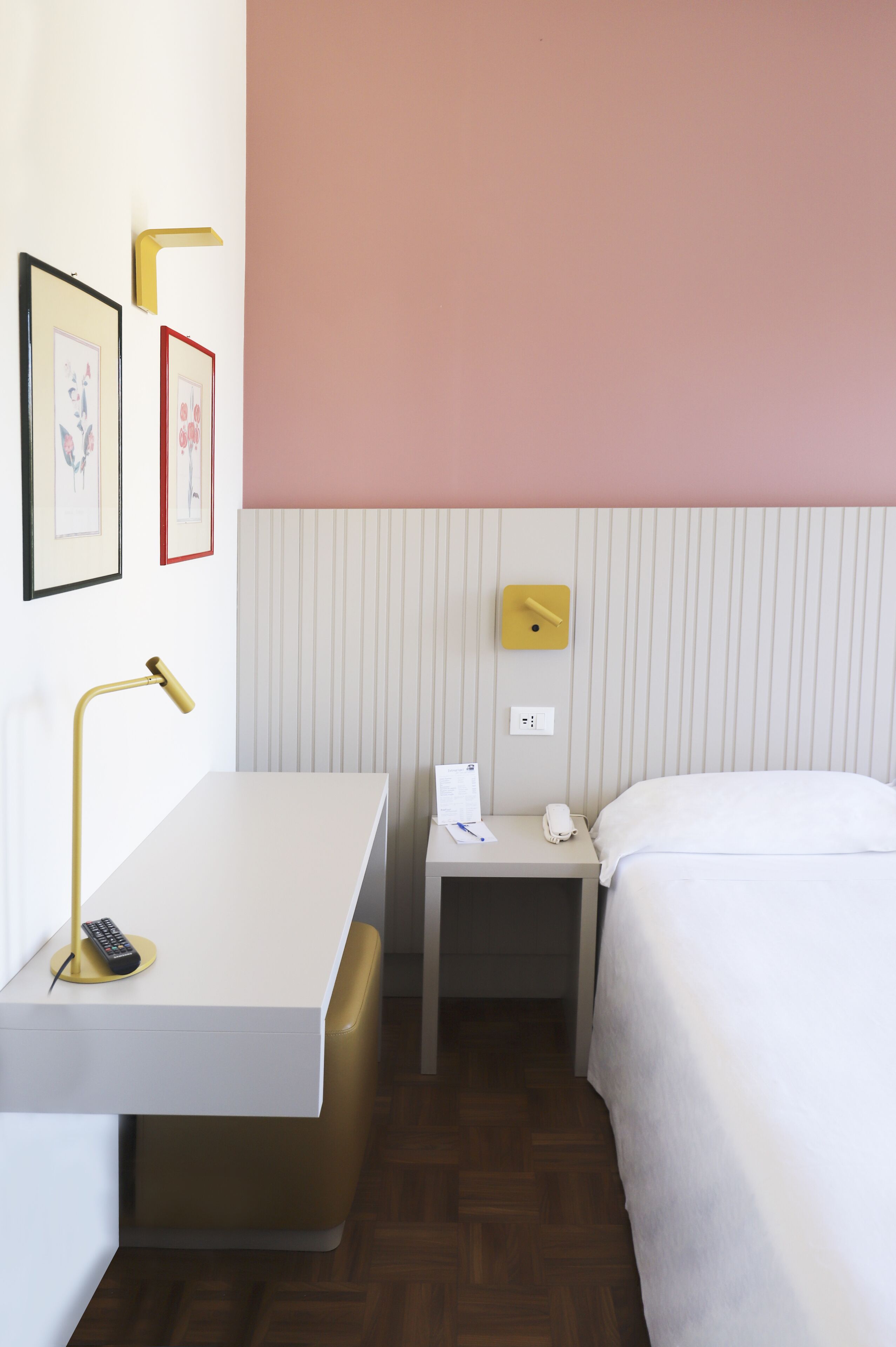 A hotel room with a white bed, a bedside table with a lamp, a nightstand with a phone, and a wall-mounted clock.