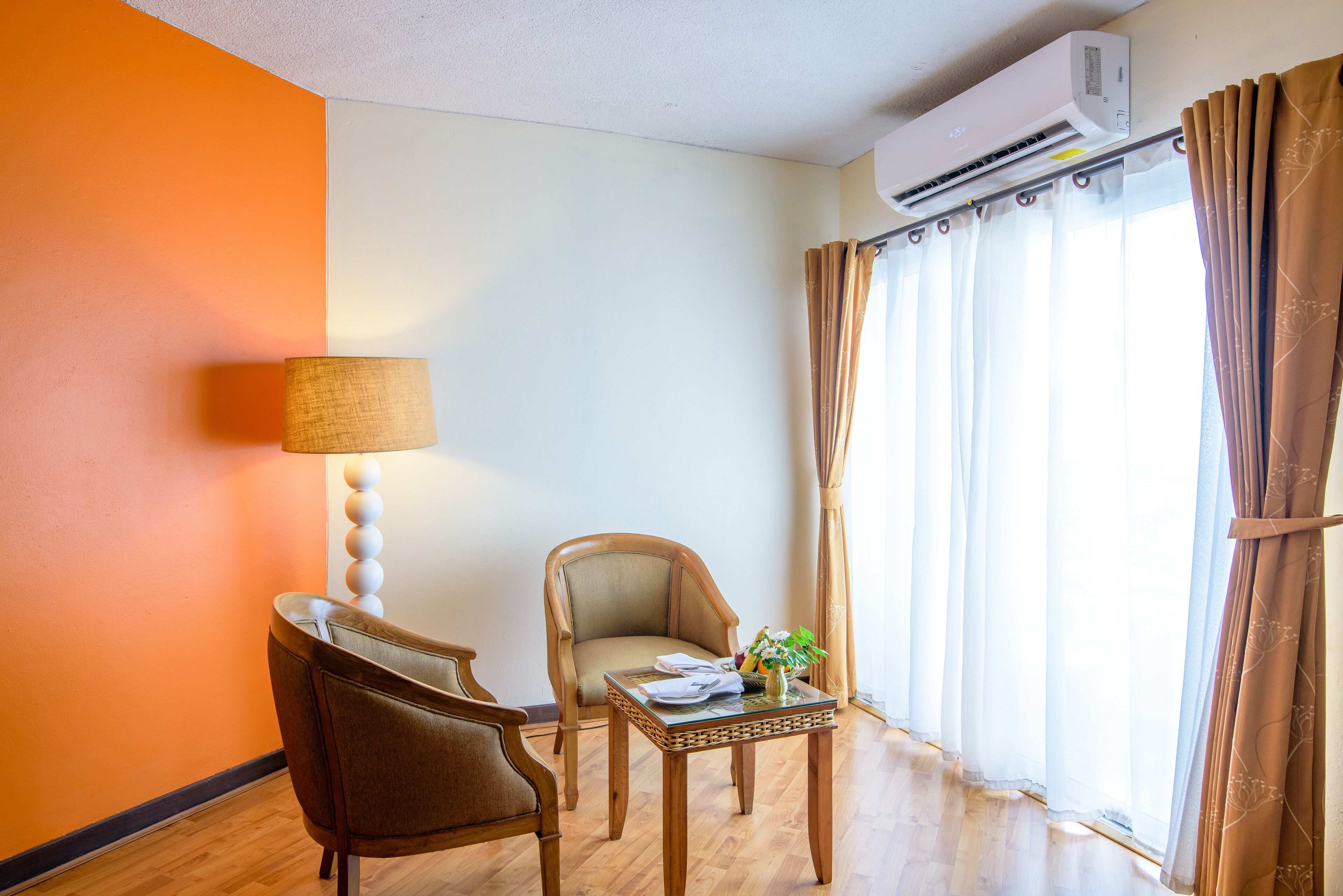 A room with a wooden floor, a brown armchair, a small table with a lamp, and a window with curtains.