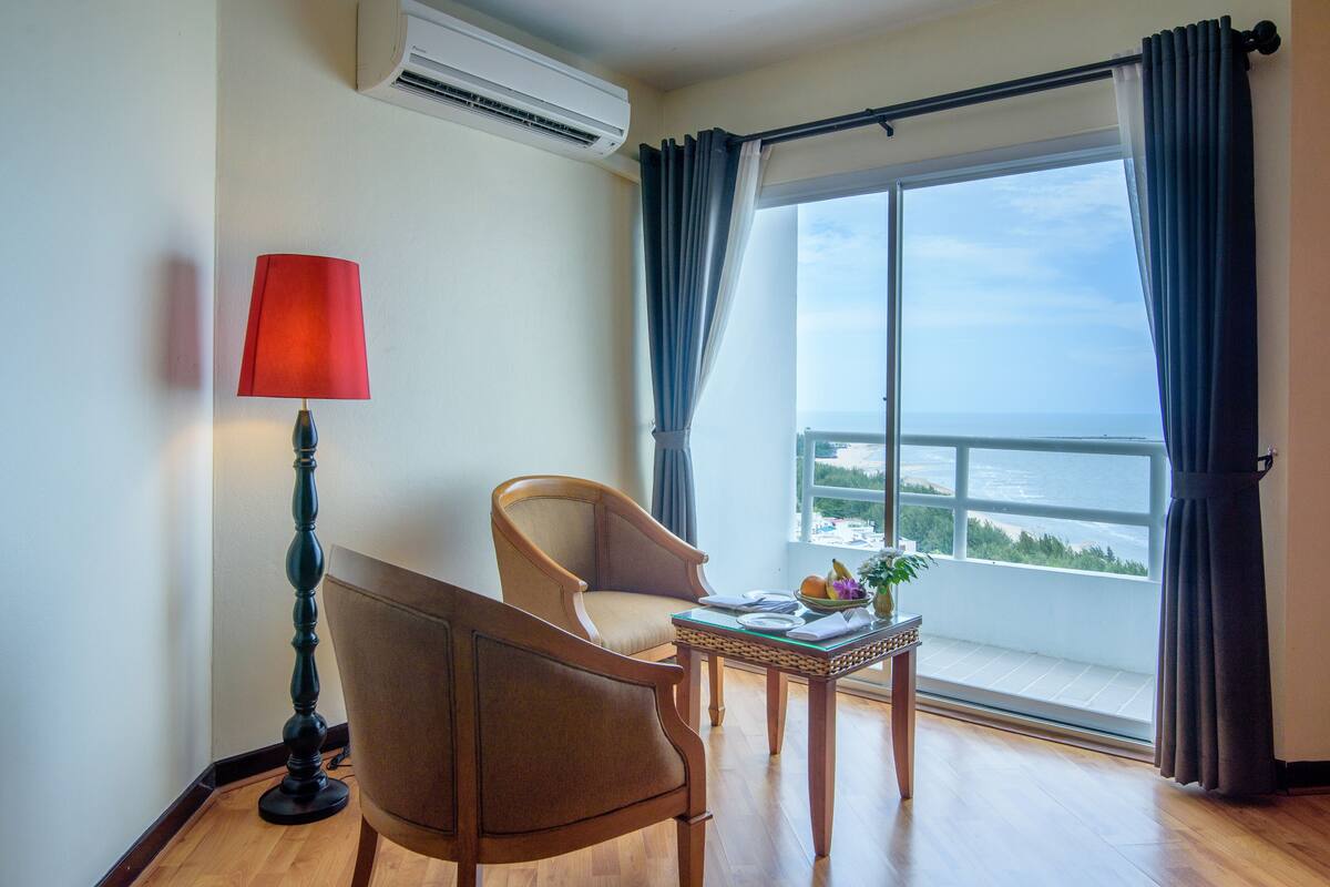 Een kamer met een stoel, een tafel met een schaal fruit, een lamp en uitzicht op zee door een groot raam.