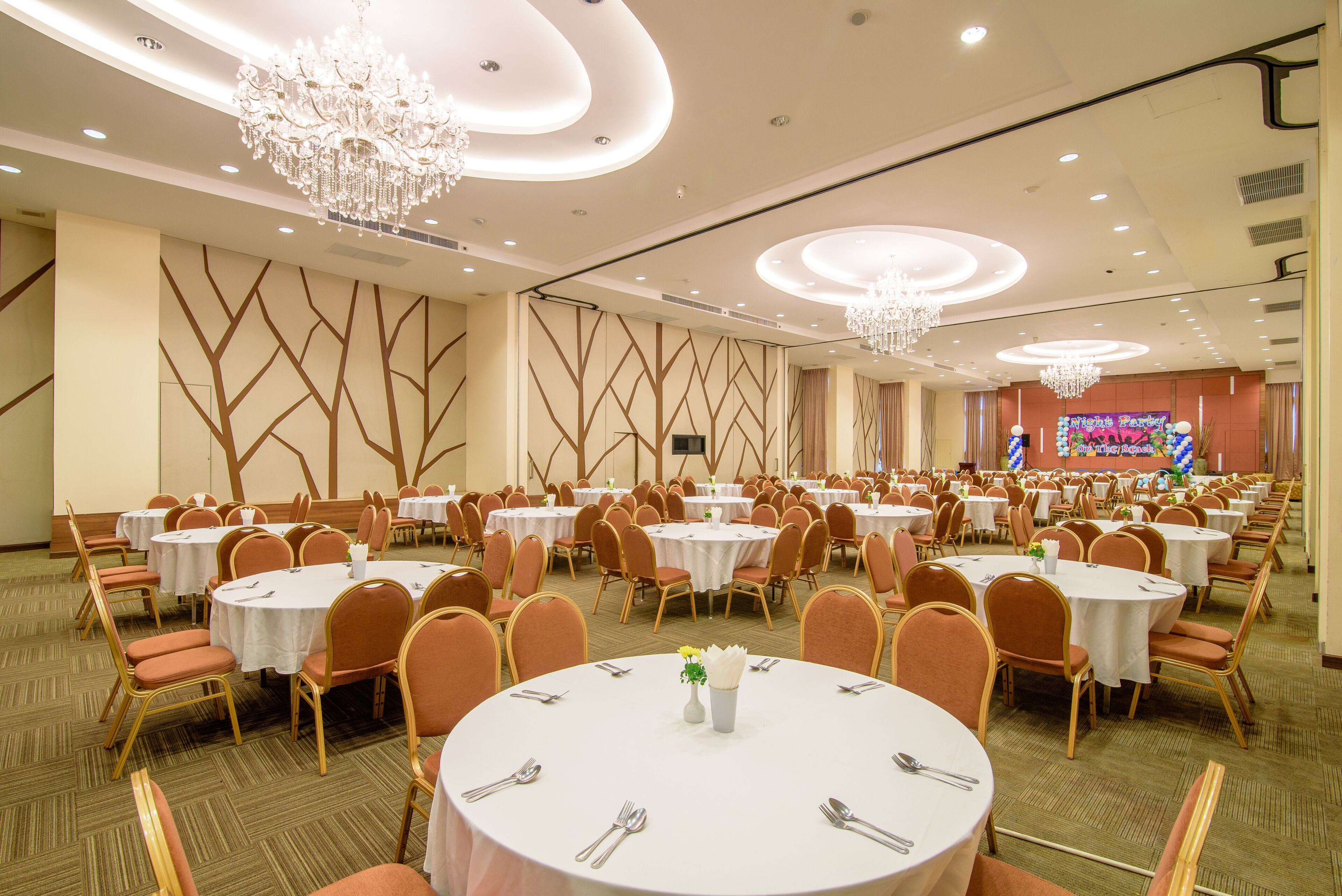 A banquet hall with round tables, chairs, chandeliers, and a stage with a colorful backdrop.