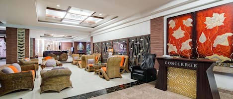 Un hall con mobiliario de mimbre, un mostrador de recepción con la etiqueta 'CONCIERGE' y una gran obra de arte floral en la pared.