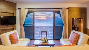 A room with a large window, a flat-screen TV, two white sofas, a small table with a flower arrangement, and a balcony with chairs. - Golden Beach Cha-Am Hotel (Cha-am)