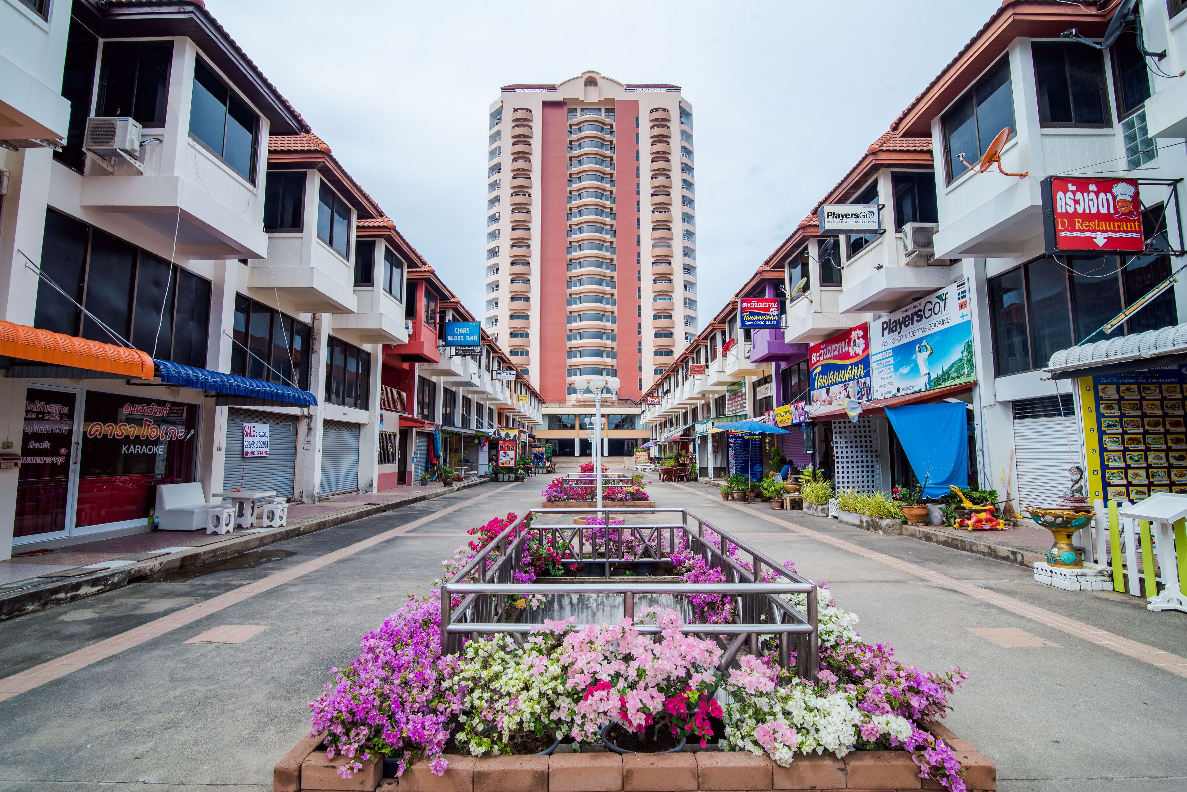 A street with a central flower bed, buildings on either side, and a tall building in the background.