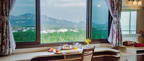 Una habitación con un ventanal grande que ofrece vistas a montañas y vegetación. Hay una bandeja de desayuno en el alféizar, una silla y un escritorio con cajones.