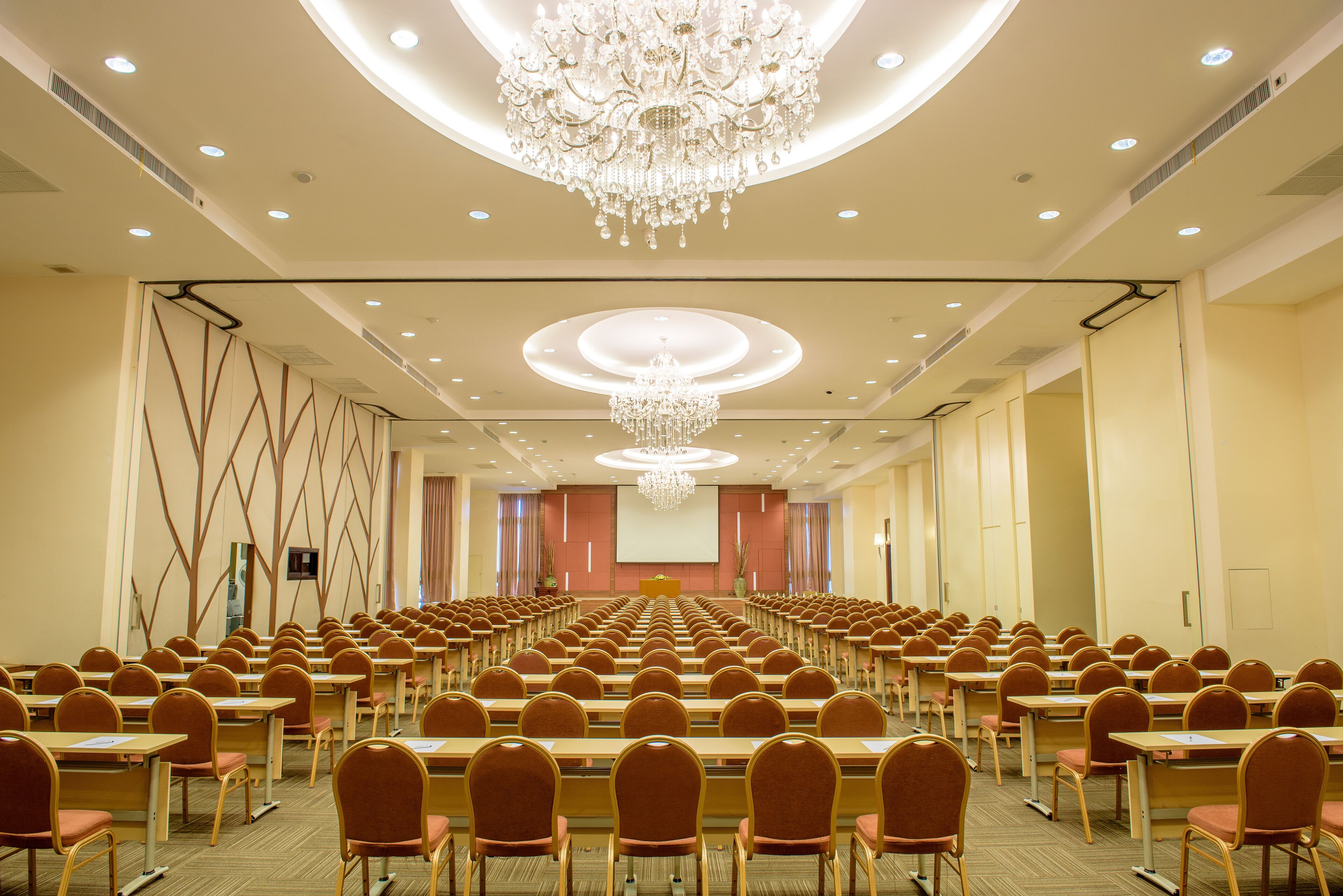 A conference room with rows of tables and chairs, a large screen, and a chandelier.