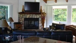 A living room with a stone fireplace, a flat-screen TV, a blue sofa, and a ceiling fan. - Bamboo Valley Inn (Haiku)