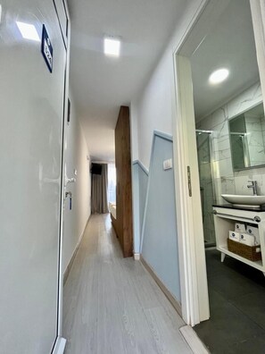 Corredor de hotel com porta de madeira, banheiro com pia e espelho, e piso em tom claro.