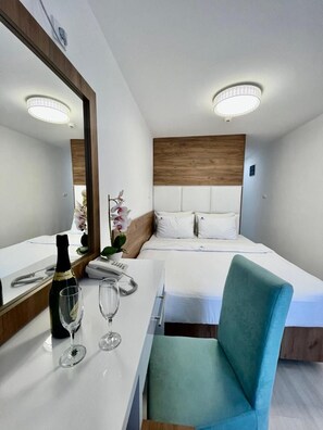 Quarto de hotel com cama, escrivaninha, garrafa de champanhe, dois copos, cadeira e um espelho grande.