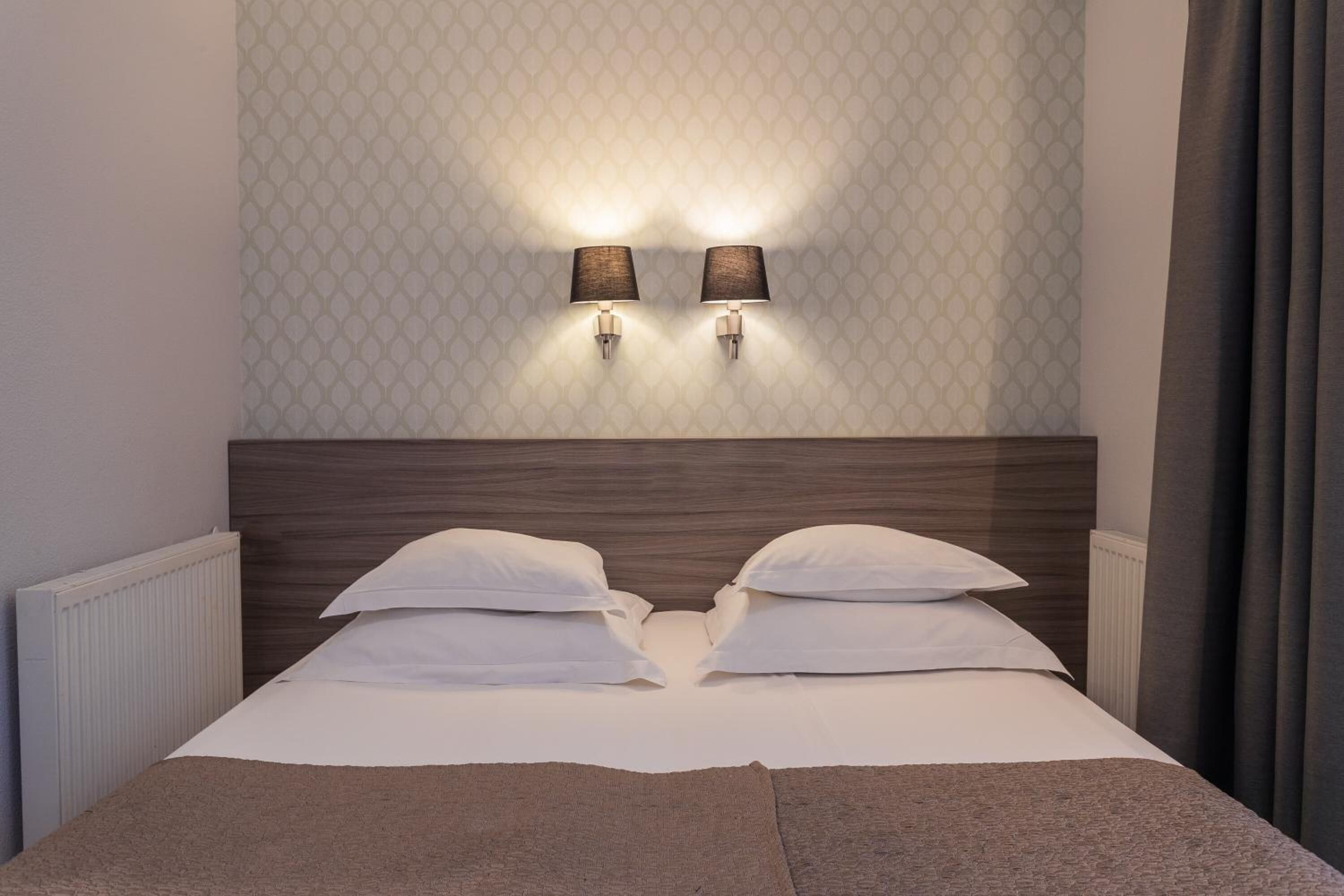 Una cama bien tendida con sábanas blancas y dos almohadas, un cabecero de madera y dos lámparas fijadas a la pared.