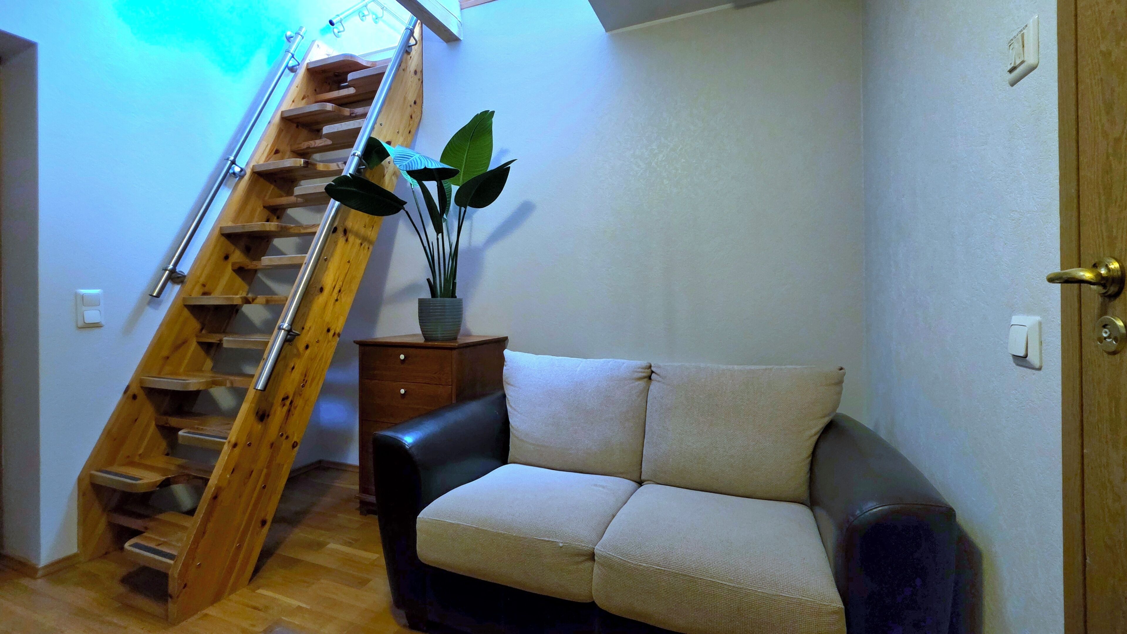 A wooden staircase, a sofa, a small wooden cabinet, and a potted plant in a room with white walls.
