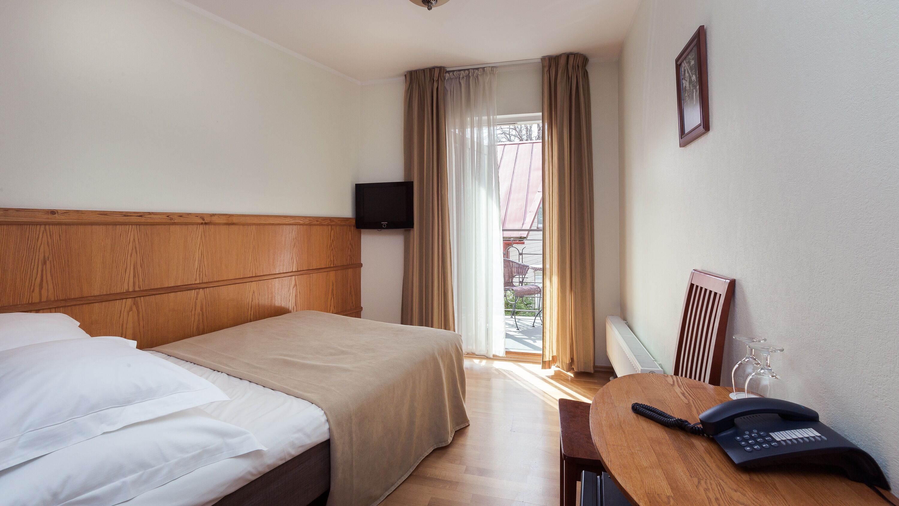 Una habitación de hotel con cabecera de madera, cama con ropa de cama blanca y beige, escritorio con teléfono y ventana con cortinas.