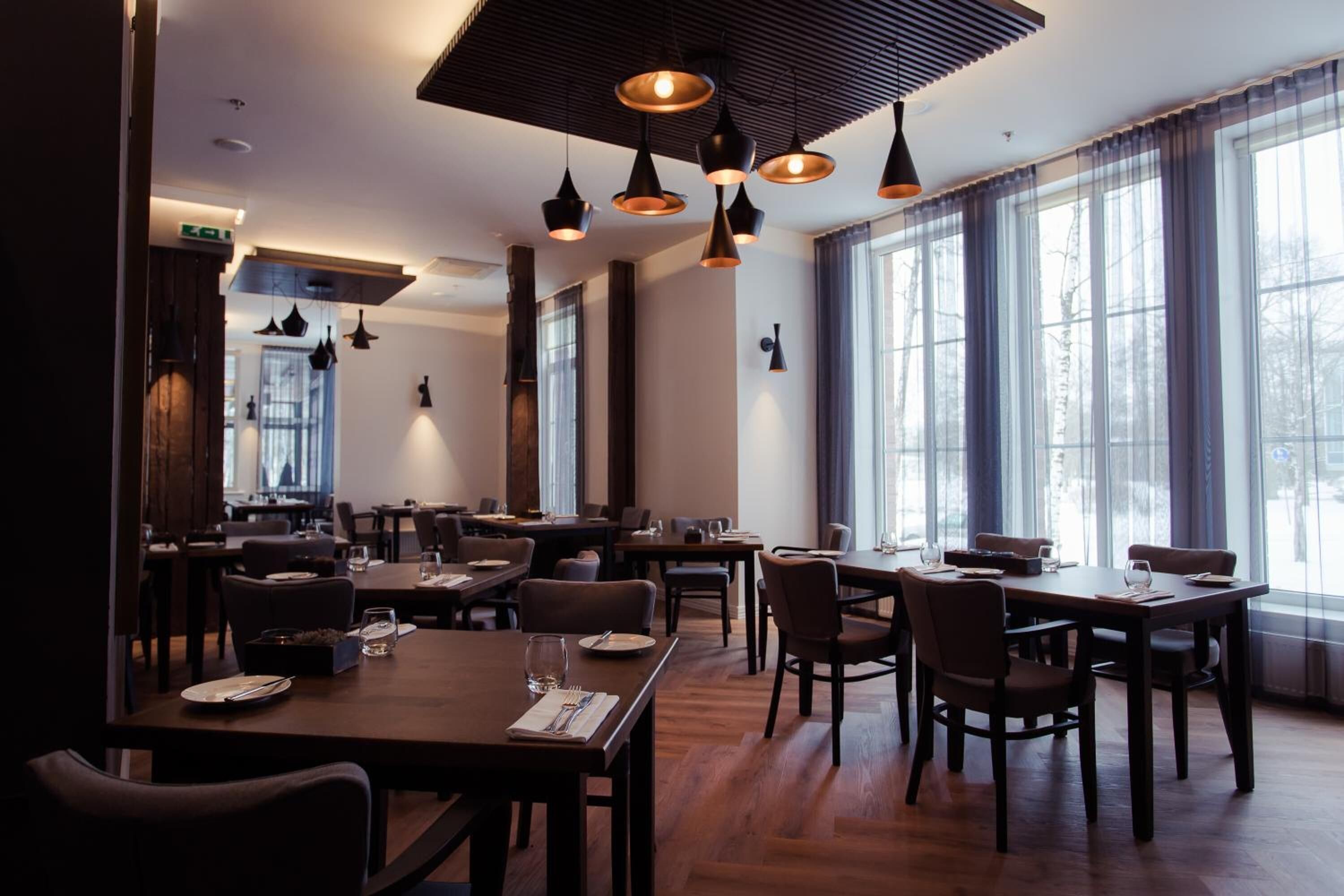 A modern restaurant with wooden tables and chairs, pendant lighting, and large windows offering a view of the outdoors.