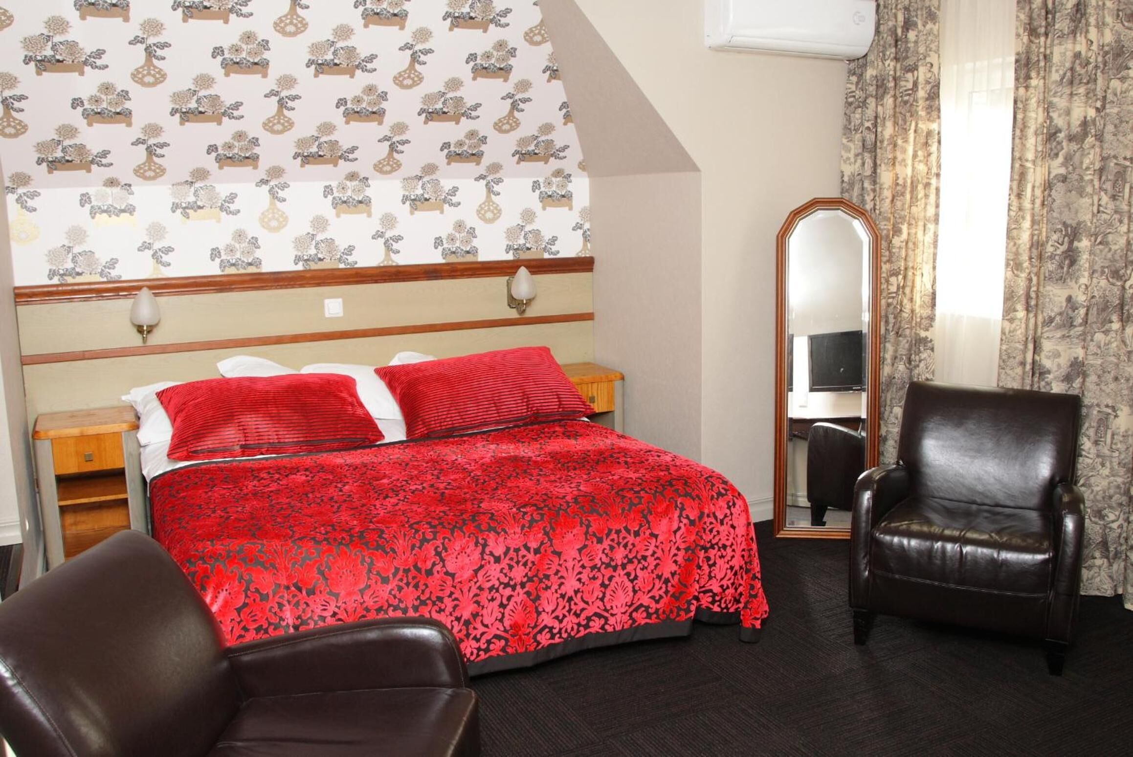 A bedroom with a bed, two red pillows, a wooden headboard, a mirror, a brown leather chair, and a patterned wallpaper.