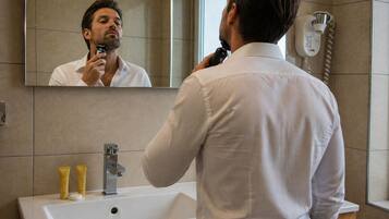 A man in a white shirt using a hair dryer in a bathroom with a sink, mirror, and a phone on the wall.