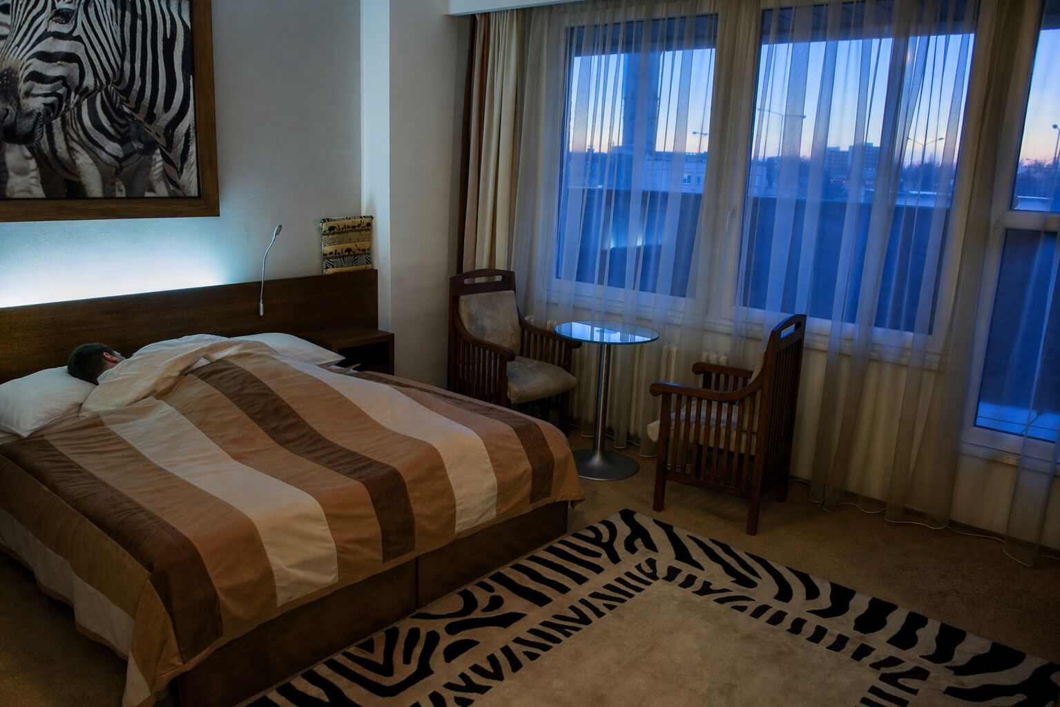 A hotel room with a bed, a chair, a small table, and a zebra-patterned rug.