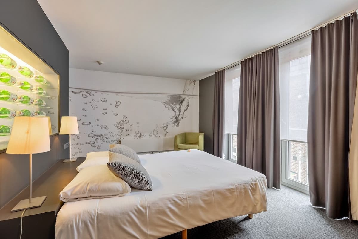 A hotel room with a large bed, a desk with a lamp, a chair, a wall with a water feature, and a window with curtains.
