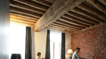 Et soveværelse med træloft, murstensvæg, seng med sort og hvidt sengetøj, skrivebord med lampe og et vindue med gardiner.