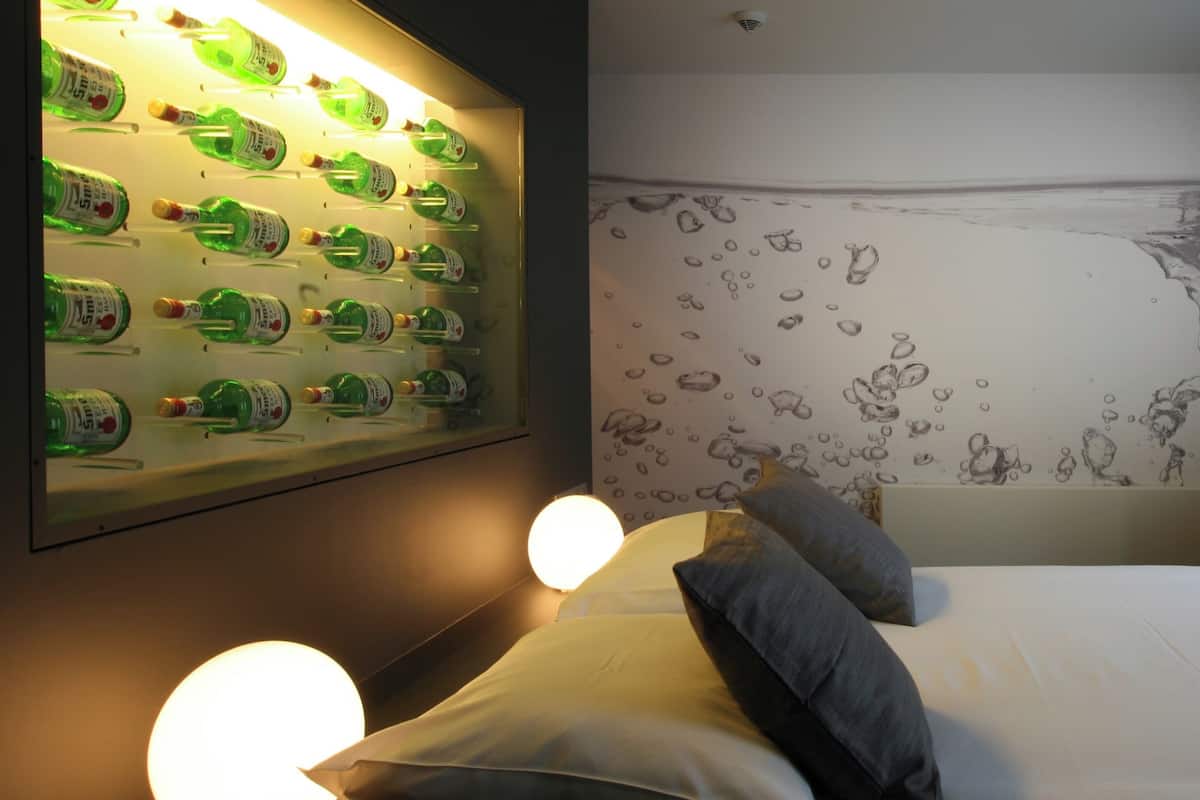 A bedroom with a wall-mounted bottle display, a bed with pillows, and a wall mural.