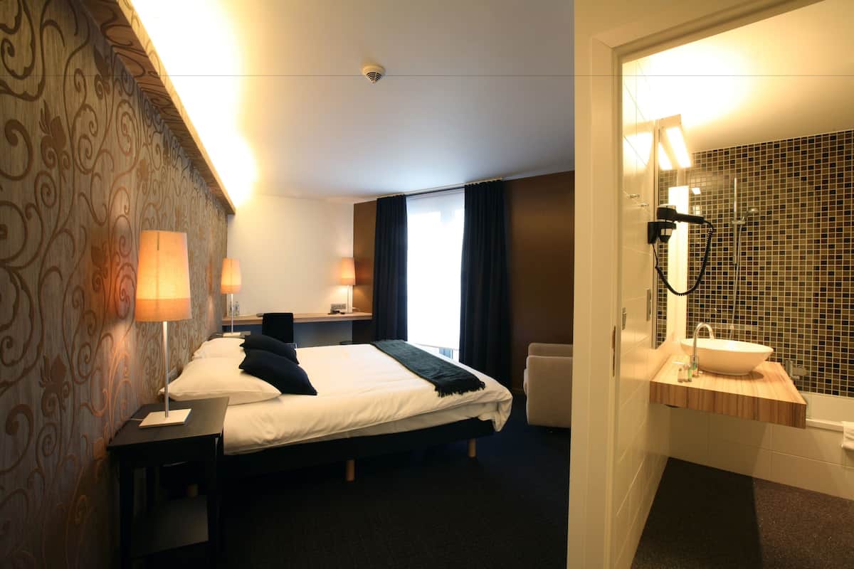 A hotel room with a bed, bedside lamps, a desk, a chair, a bathroom with a sink and mirror.