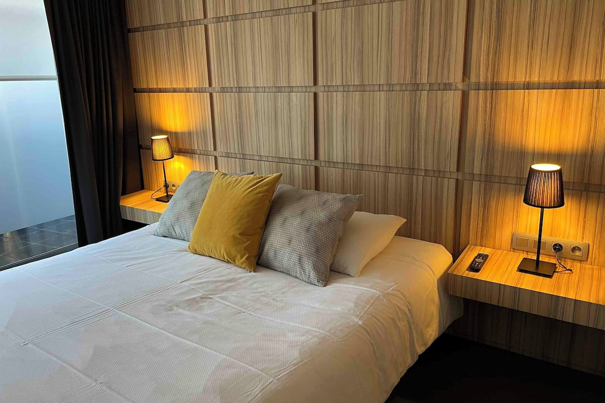 A hotel room with a large bed, wooden headboard, two bedside tables with lamps, and a view of the outside.