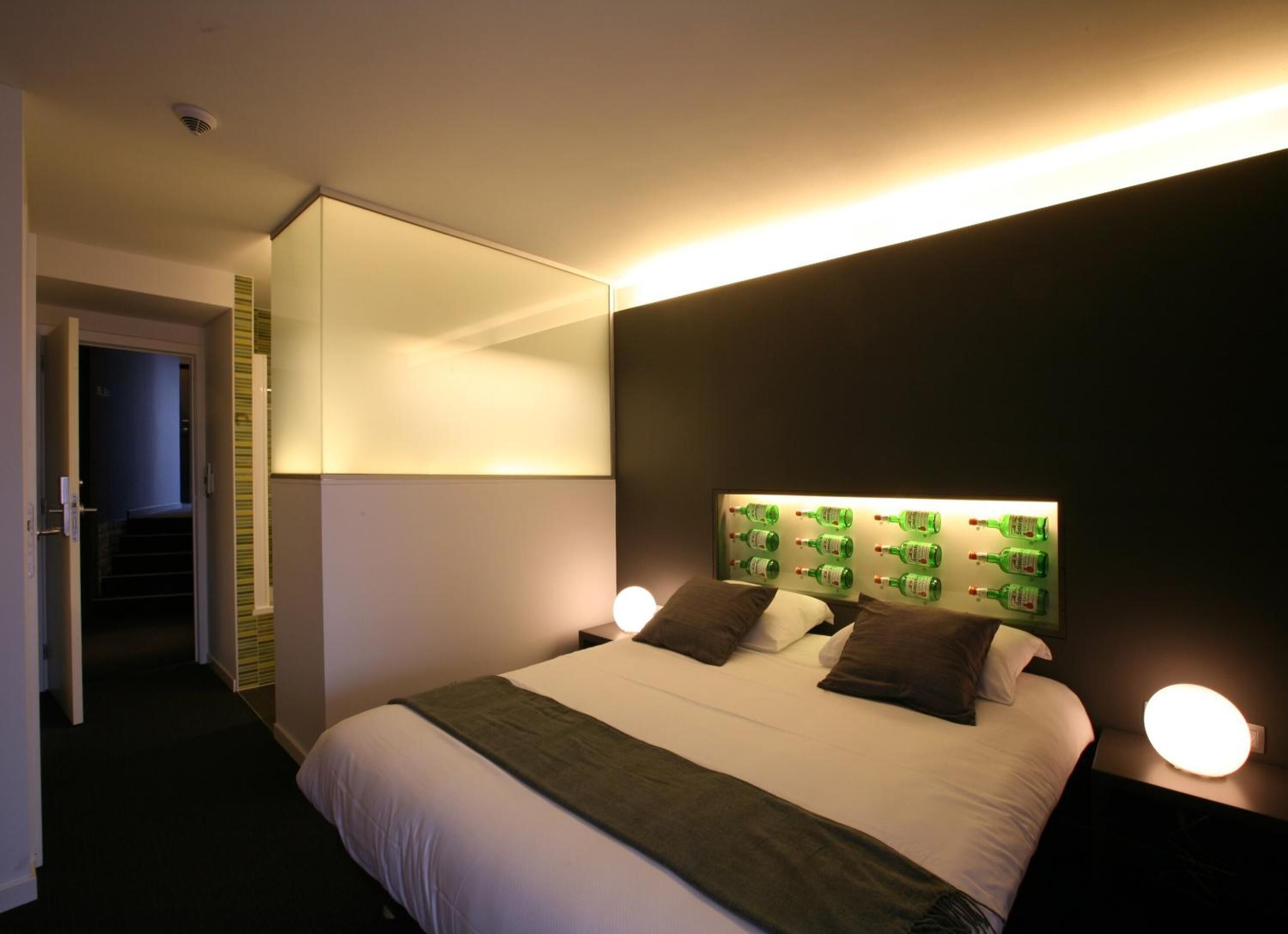 A hotel room with a large bed, a mirror above the bed, a headboard with green bottles, and a staircase leading to an upper level.