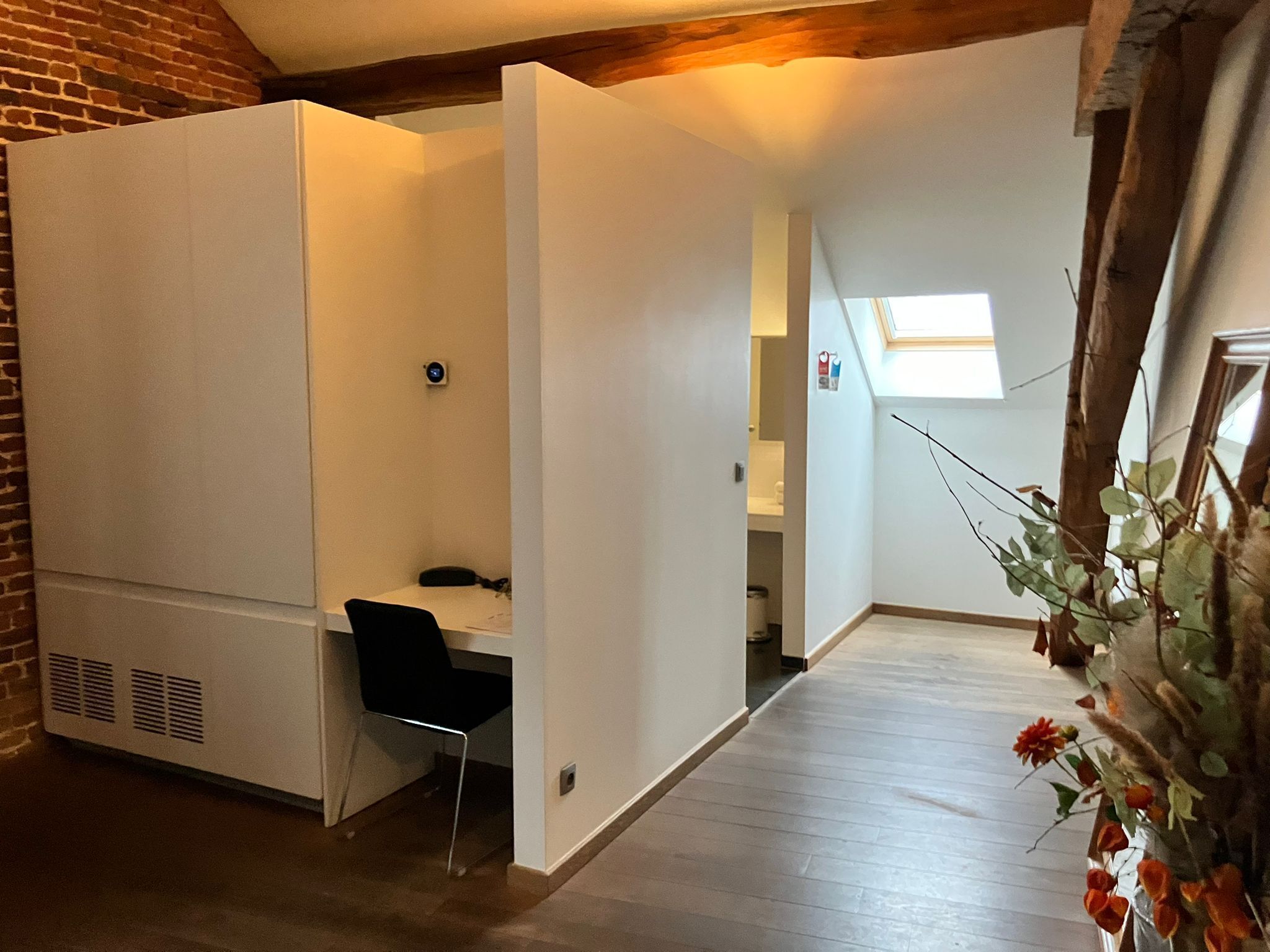 A room with a desk, chair, and a small window. There is a brick wall on one side and a white partition on the other.