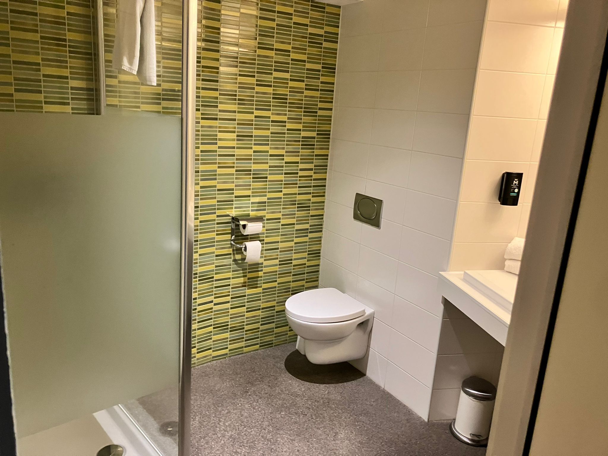 Et badeværelse med glasvægs brusebad, toilet og håndvask med sæbedispenser.