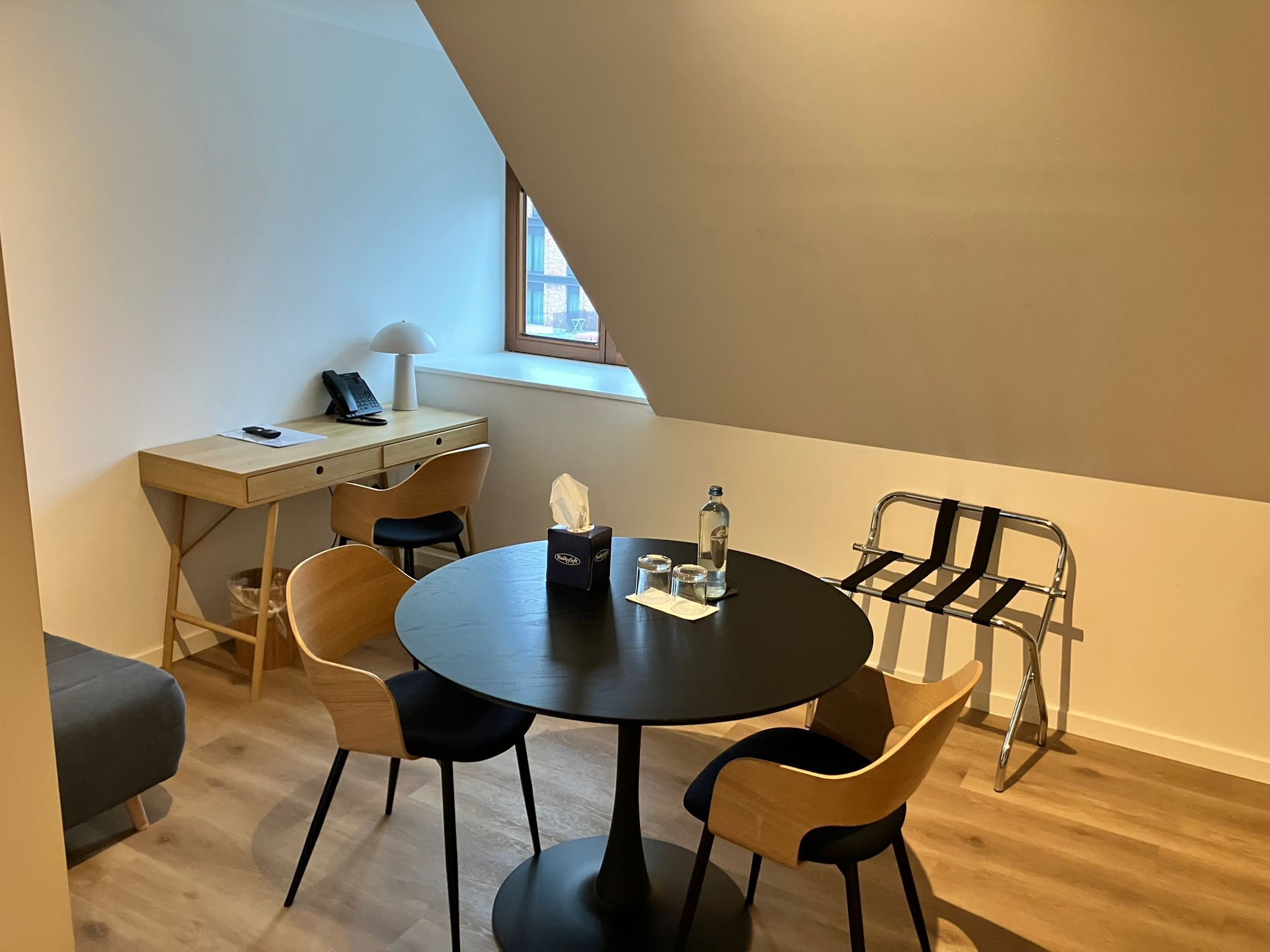 A small, modern room with a round table, four chairs, a desk with a lamp, a chair, and a window.
