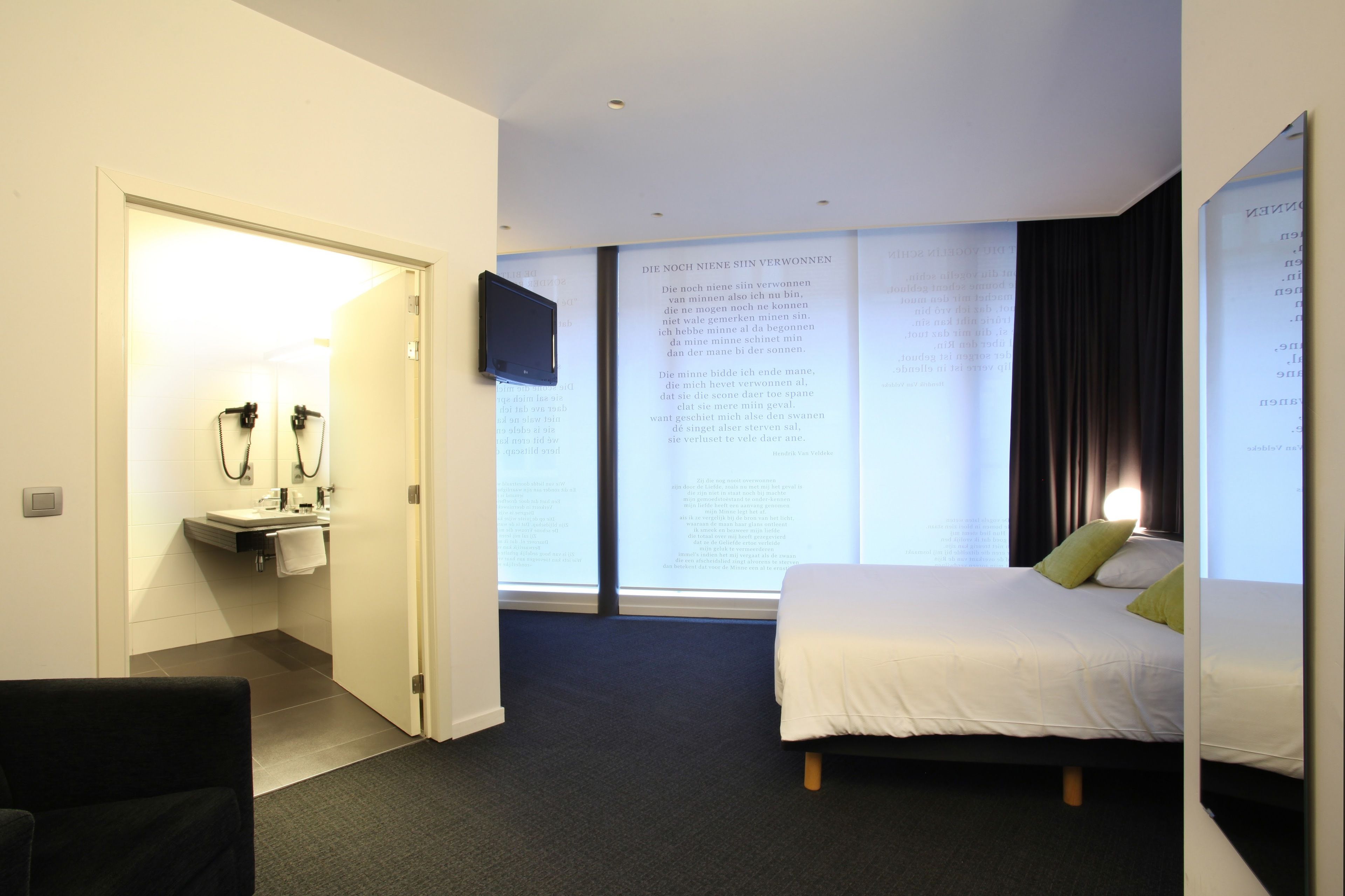 Habitación de hotel con cama, televisión, baño con lavamanos y espejo, y una ventana grande con cortinas.