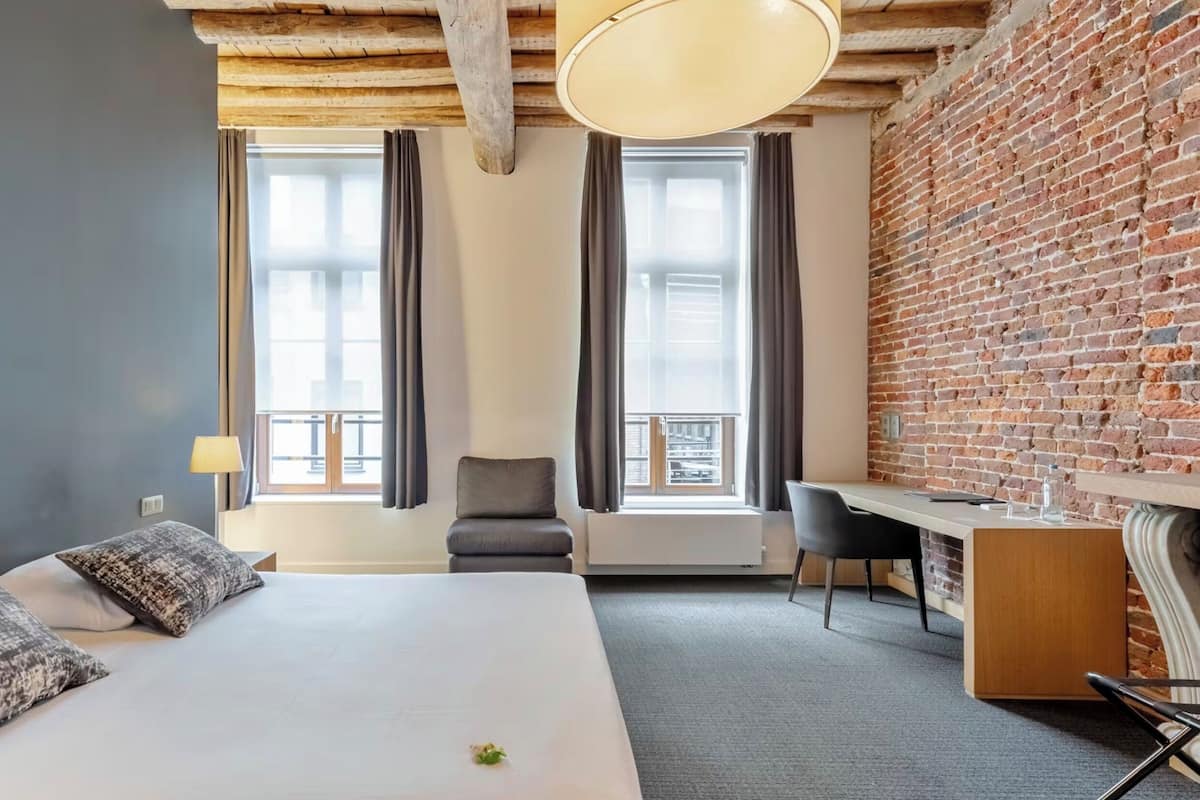 A hotel room with a large bed, a desk, a chair, and a view of the city through large windows.