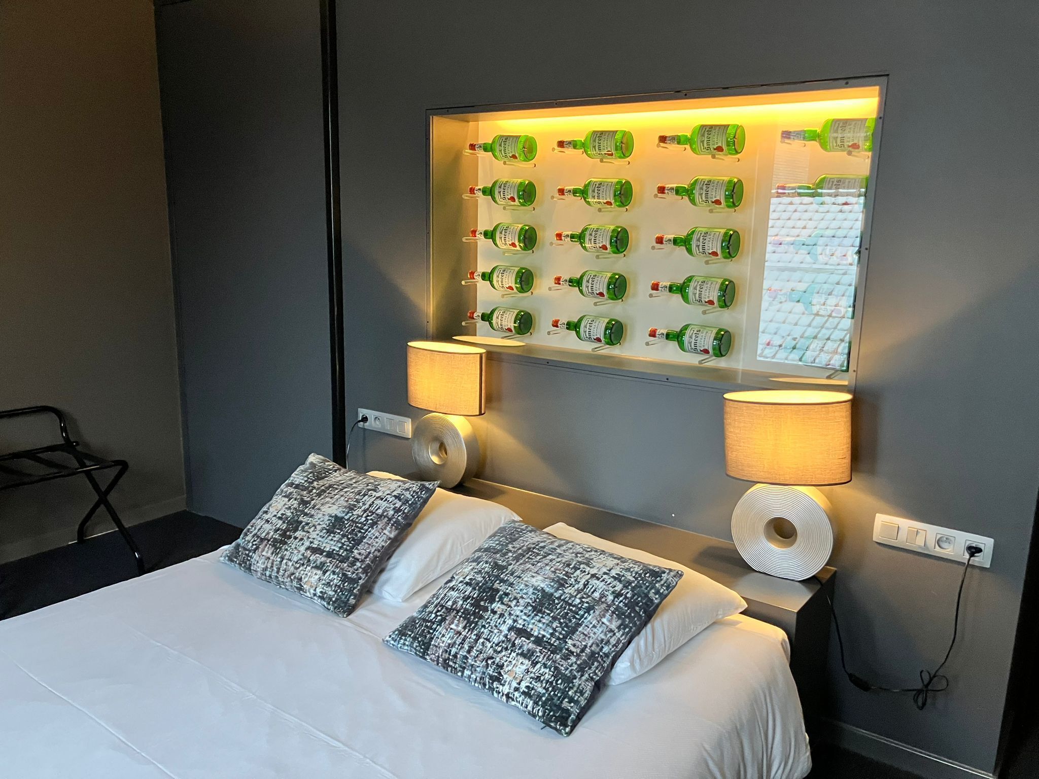 Ein Hotelzimmer mit einem Bett, zwei gemusterten Kissen, einer Wandablage mit grünen Flaschen, zwei Wandlampen und einer Steckdose.