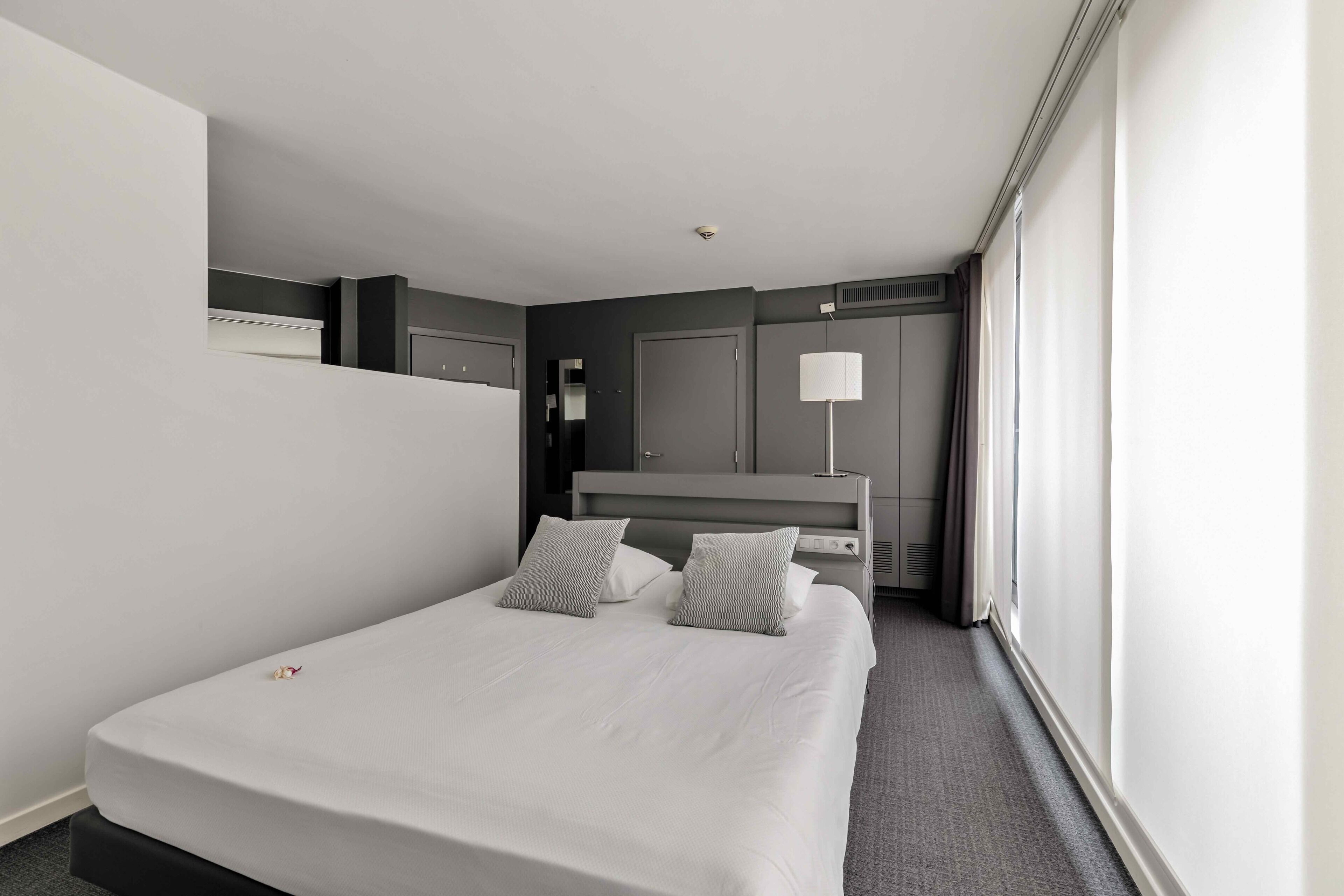 A modern hotel room with a large bed, a bedside table, a lamp, and a view of the city.