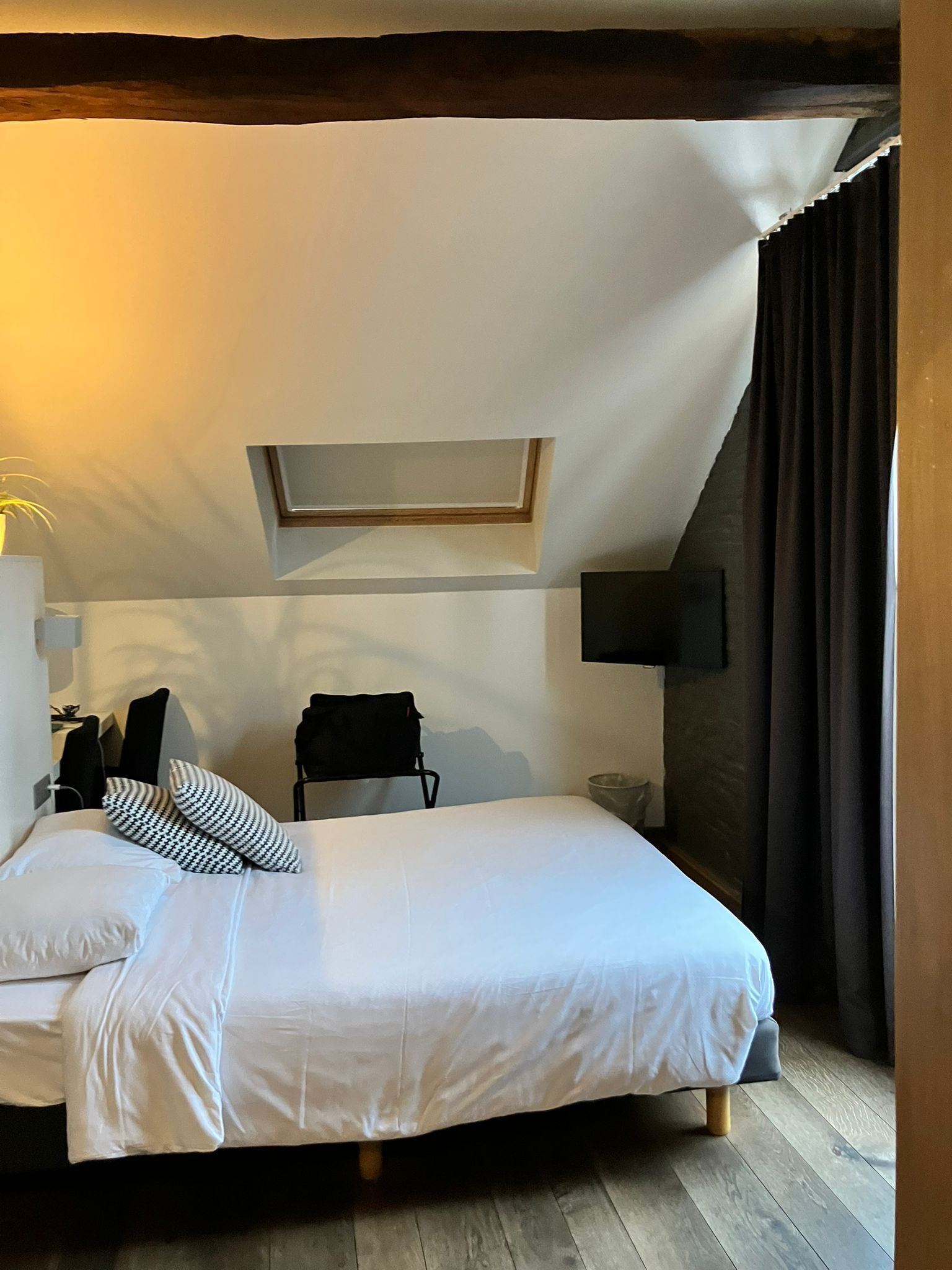 A bedroom with a bed, a small table, a chair, a wall-mounted TV, and a window with a view of the outside.