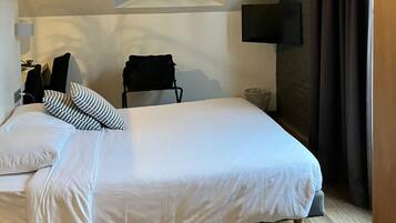 A bedroom with a bed, a small table, a chair, a wall-mounted TV, and a window with a view of the outside.