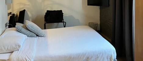 Un dormitorio con cama, una mesita, una silla, televisor montado en la pared y ventana con vistas al exterior.