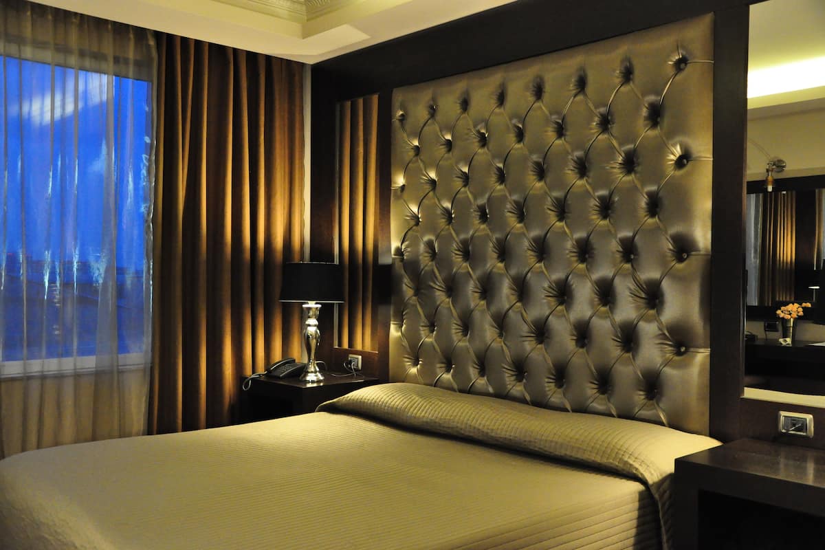 A hotel room with a large bed, a tufted headboard, a bedside lamp, and a view of the outdoors through a window with curtains.