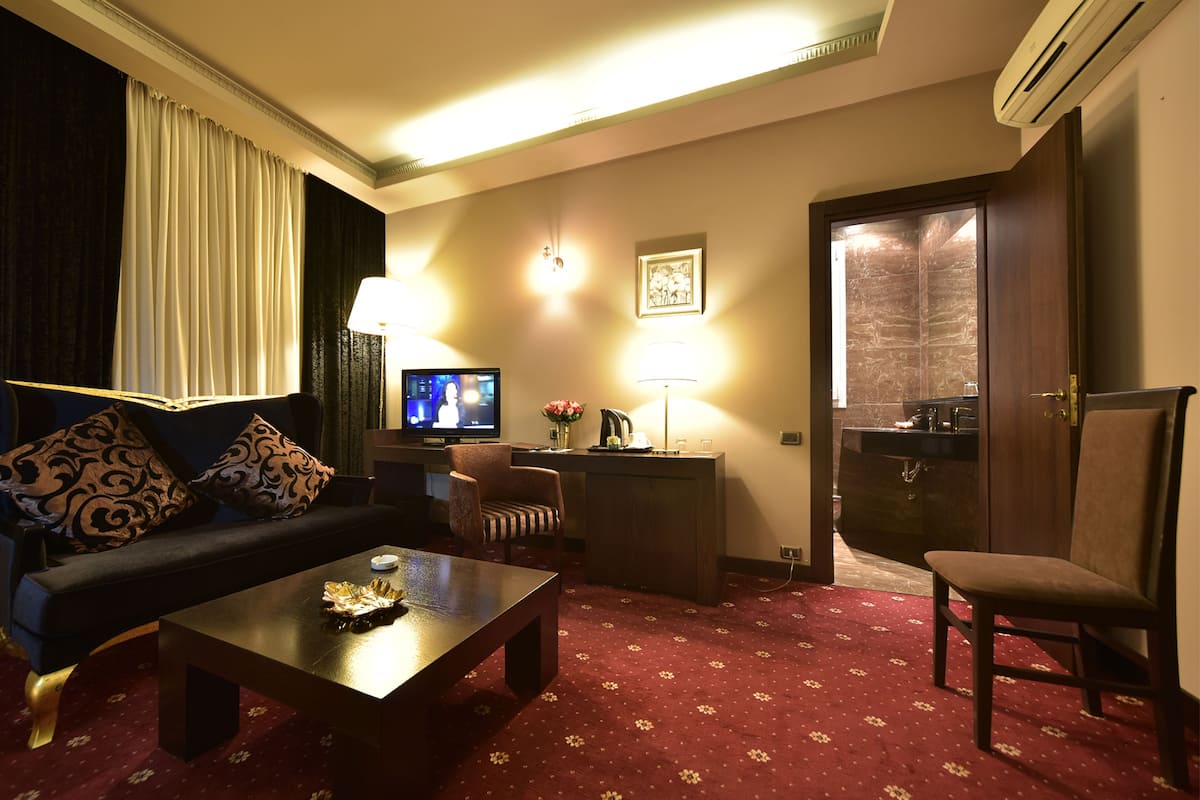 A hotel room with a sofa, armchair, coffee table, TV, desk, lamp, and an air conditioning unit.