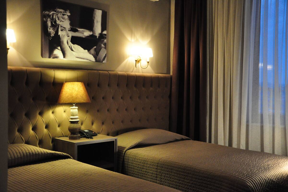 A hotel room with a tufted headboard, a bedside lamp, a framed picture on the wall, and a view through sheer curtains.