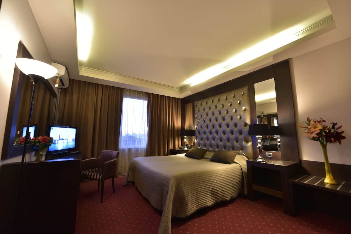 A hotel room with a large bed, a desk with a computer, a chair, a bedside table with a lamp, a mirror, and a vase with flowers.