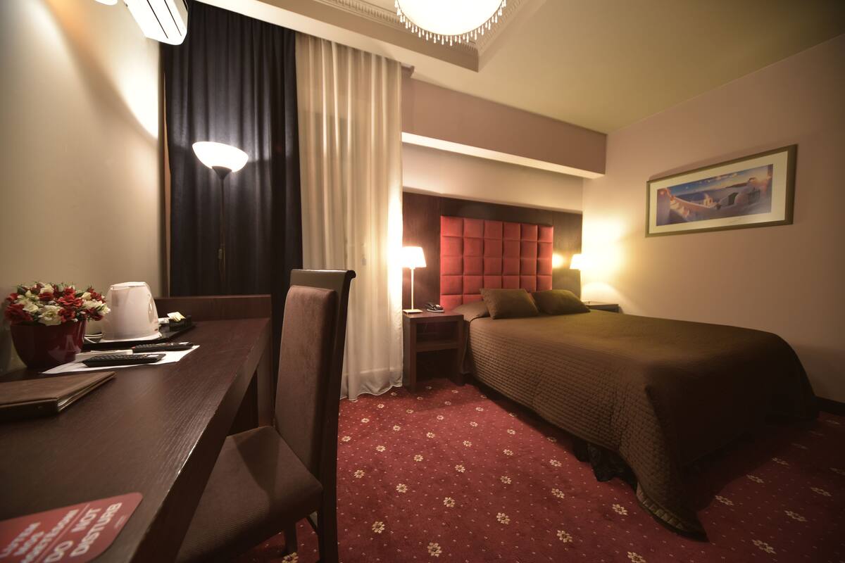 A hotel room with a bed, a desk with a lamp, a chair, a nightstand, and a painting on the wall.