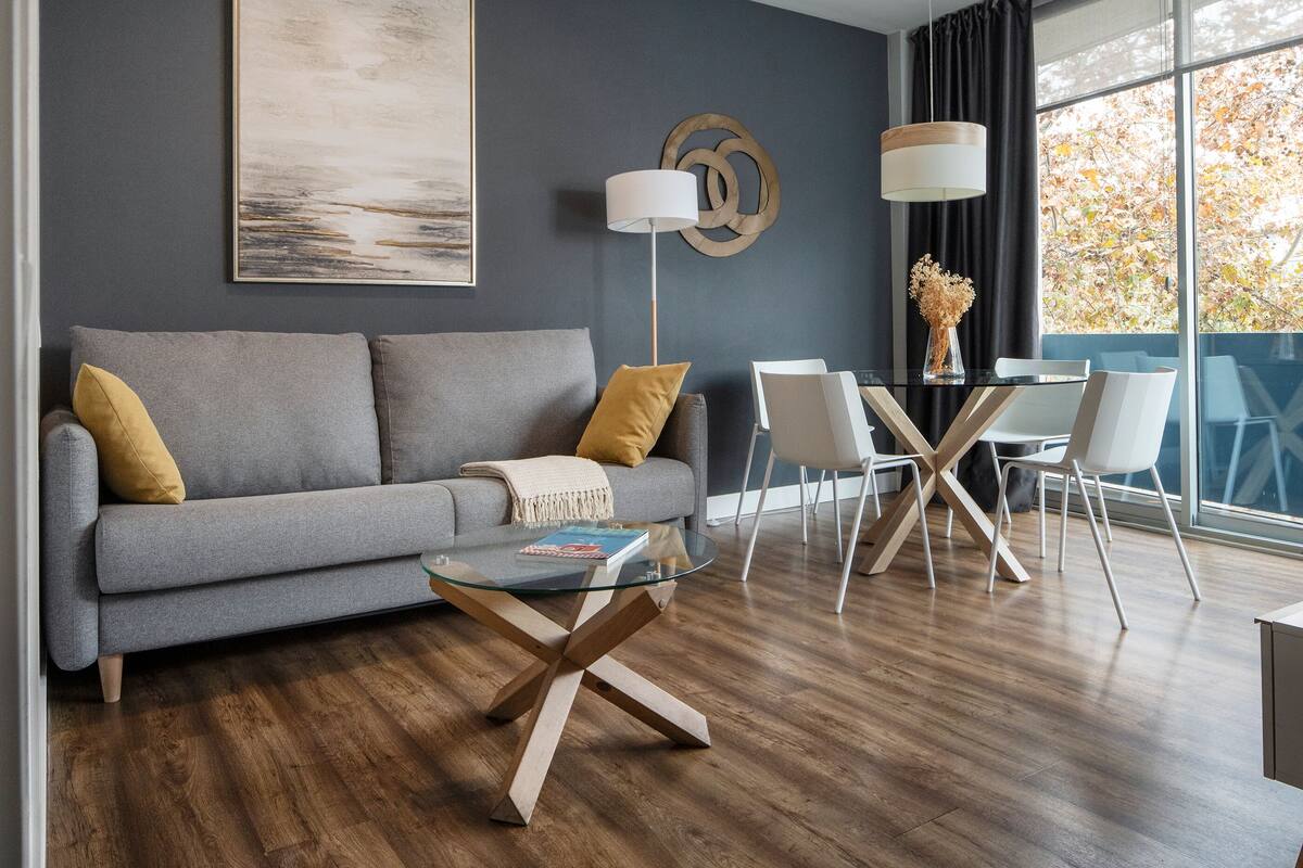 A modern living room with a grey sofa, a glass coffee table, and a dining area with white chairs and a glass table.