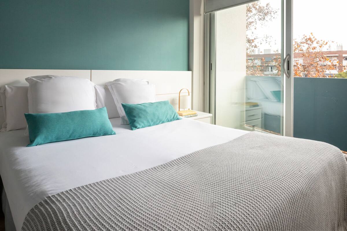 A hotel room with a bed, white bedding, teal pillows, a white headboard, a nightstand with a lamp, and a view of the city through a large window.