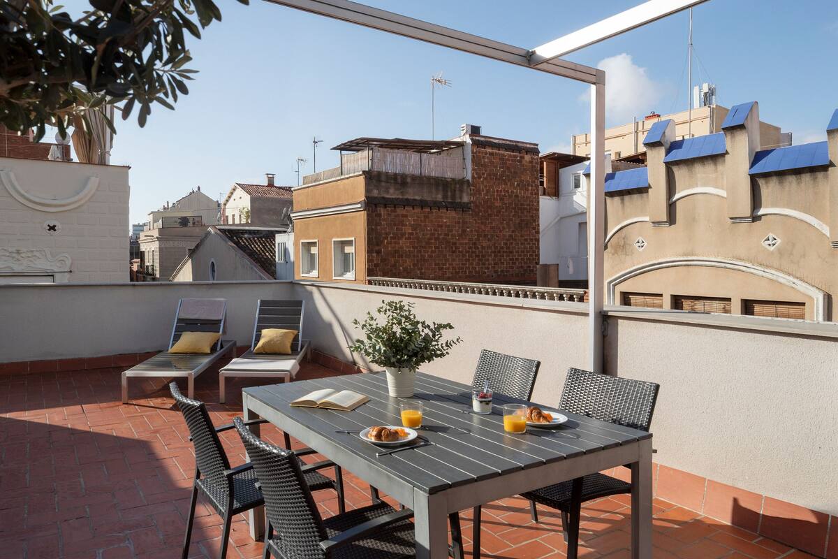 A rooftop terrace with a table set for two, surrounded by chairs, a potted plant, and a view of the cityscape.