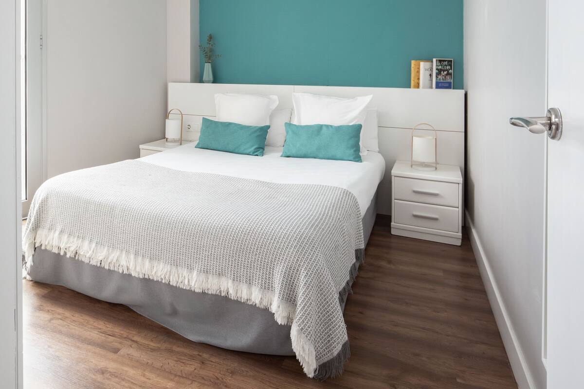 A bedroom with a bed, white nightstands, a teal accent wall, and a wooden floor.
