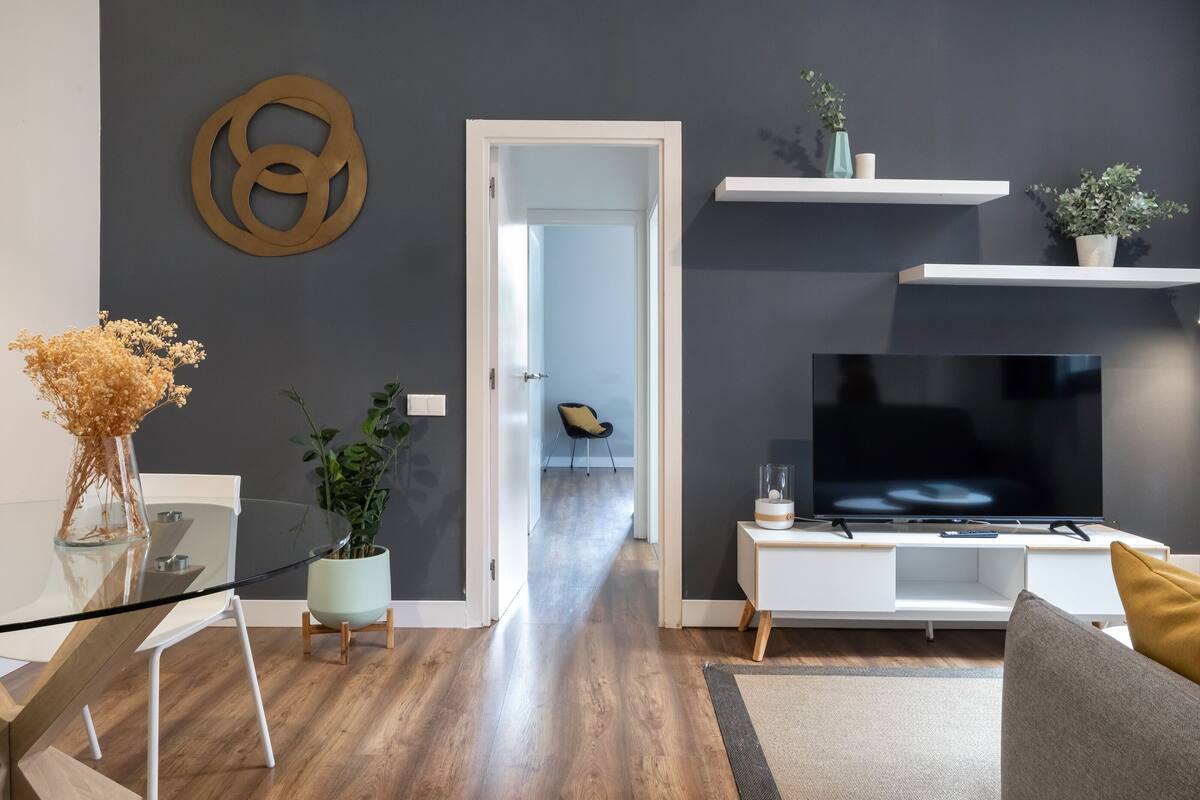 A modern living room with a flat-screen TV, a glass coffee table, and a sofa. There is a hallway leading to another room.