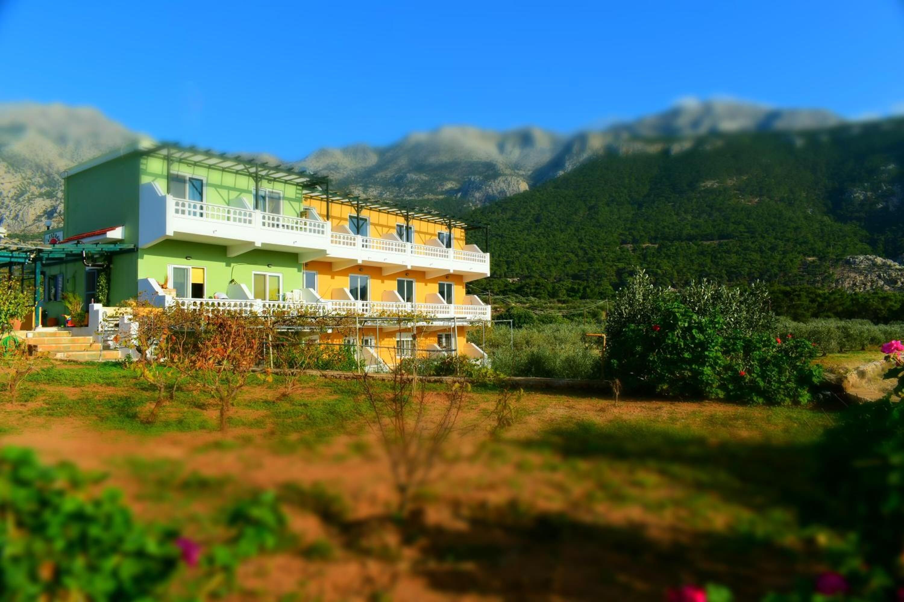 A two-story building with a balcony, surrounded by greenery and mountains in the background.